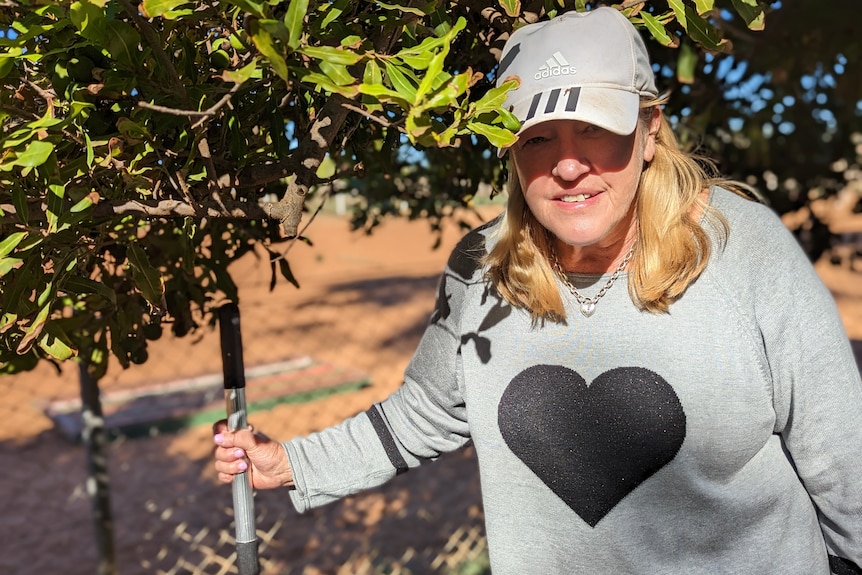 A white woman, Jan, with blonde hair smiles in a grey cap as she holds a nut picker under a macadamia tree.