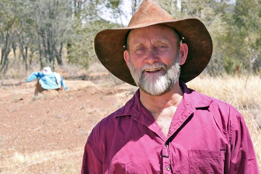 Smiling man in burgundy shirt, wide-brimmed hat stands in front of bush and termite mound dressed up as horse and rider.