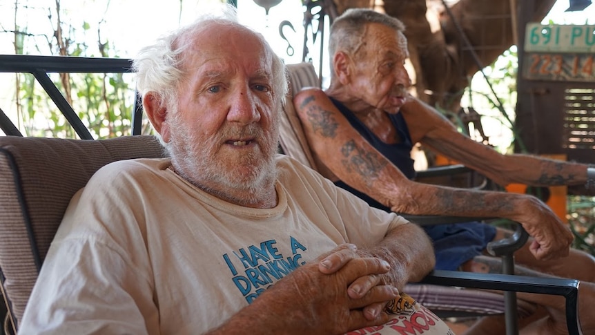 Two old men sit together on a couch in the shade.
