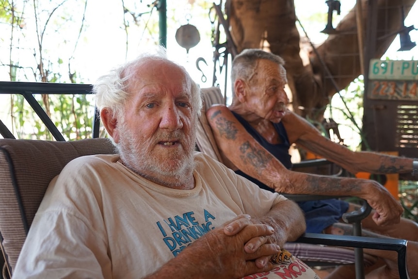 Two old men sit together on a couch in the shade.