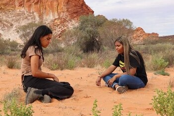 Two young women sit on red soil looking down. Behind them is a large rockface and expanding blue sky.