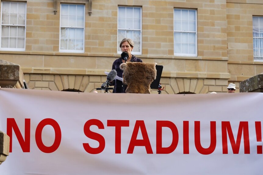 A woman stands at a lectern behind a sign which which says 'No stadium'