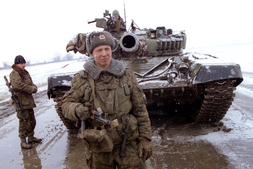 A soldier in winter uniform stands in front of a tank, with two other soldiers visible behind him