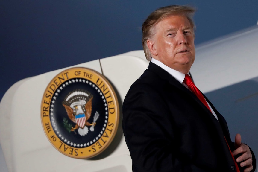 A man in a suit stands at the door to a plane bearing the seal of the president