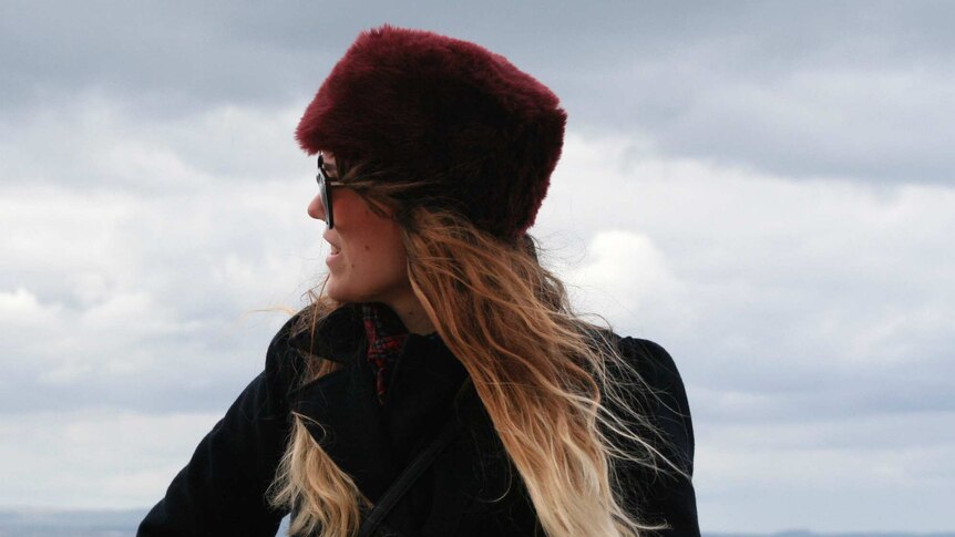 Mid-shot of a young woman in a red fur hat in side profile with grey skies in the background.
