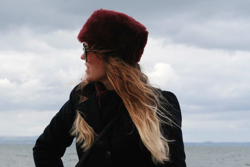 Mid-shot of a young woman in a red fur hat in side profile with grey skies in the background.