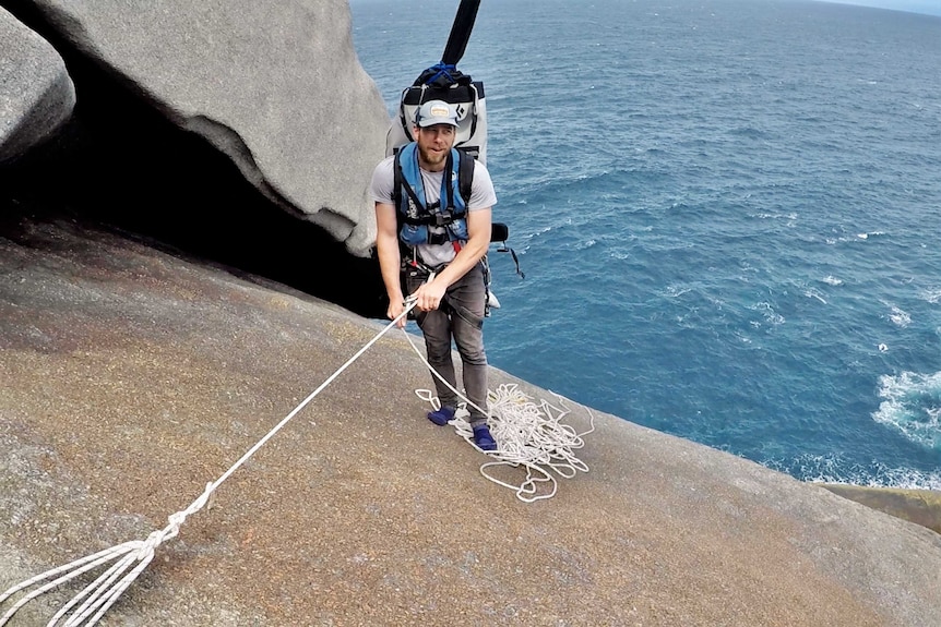 A man prepares to abseil down a cliff with the ocean behind him.