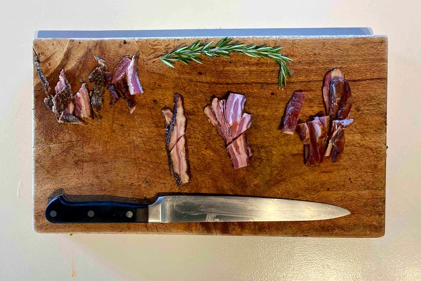 A wooden chopping board with a large knife, pieces of meat and a sprig of rosemary.