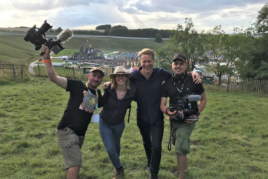Four standing with arms around each other holding camera equipment with festival in background.