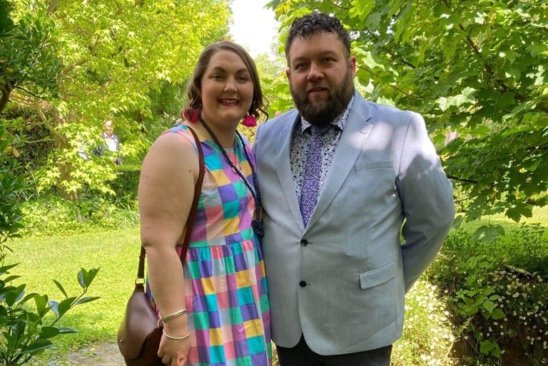 A woman dressed smartly in dress with a rainbow of squares on it and a man dressed in a grey suit stand in a lush garden.