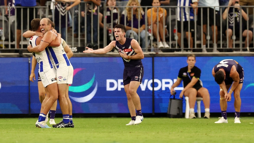 A Fremantle AFL player shouts and gesticulates as two North Melbourne teammates hug after the final siren in a game.