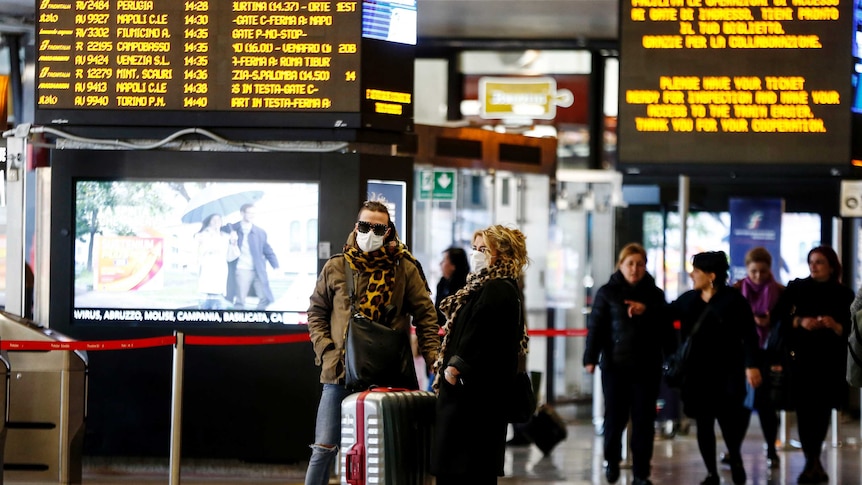 Two people wearing face masks with suitcases in front of an arrivals and departures screen.