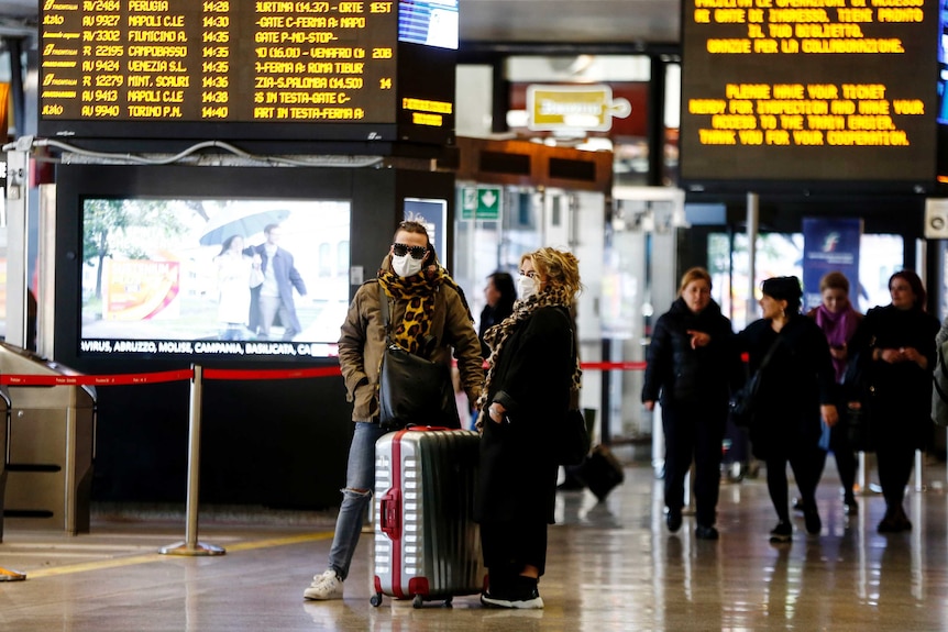 Two people wearing face masks with suitcases in front of an arrivals and departures screen.