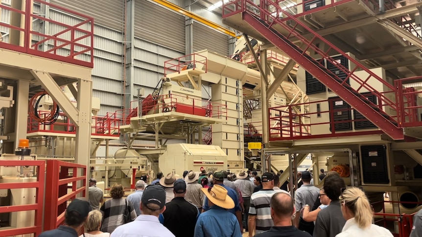 A group of people gather in a large shed between industrial machinery used for ginning cotton.