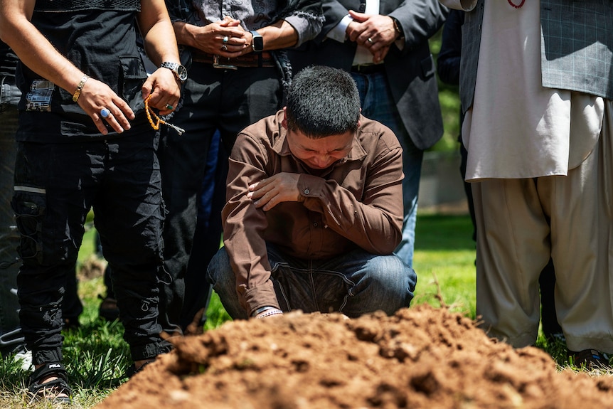 A man kneels and cries into his forearm as other men stand around him with a mound of earth in front