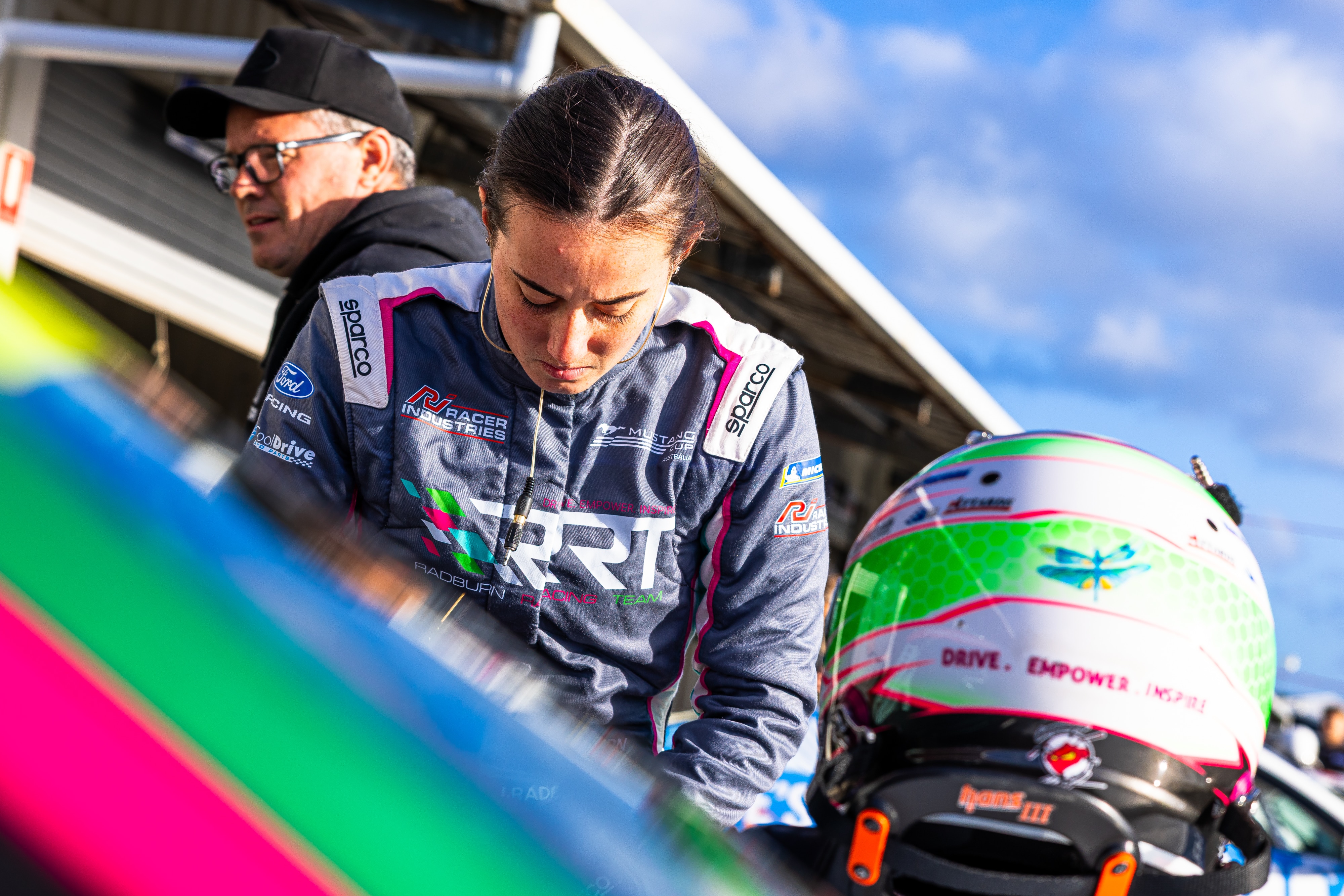 Imogen olha para seu carro com o capacete apoiado nele.