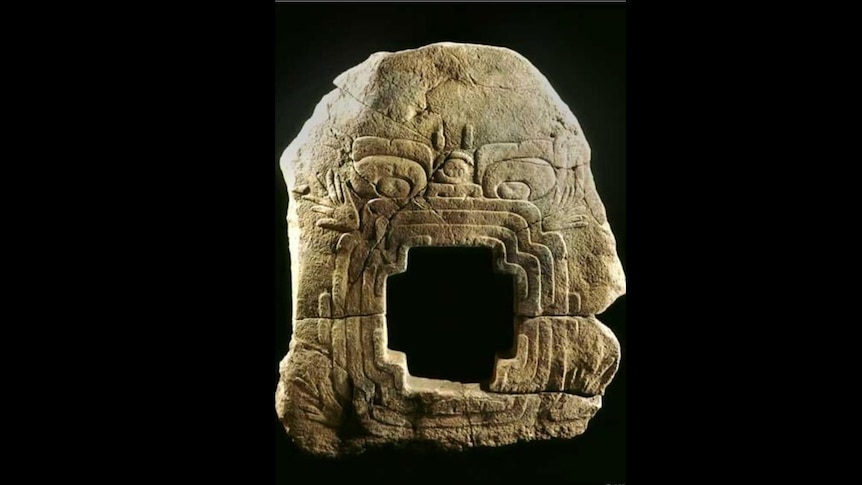 A bas-relief stone carving on a black background. It depicts a face with an open square mouth ringed by three concentric bands.