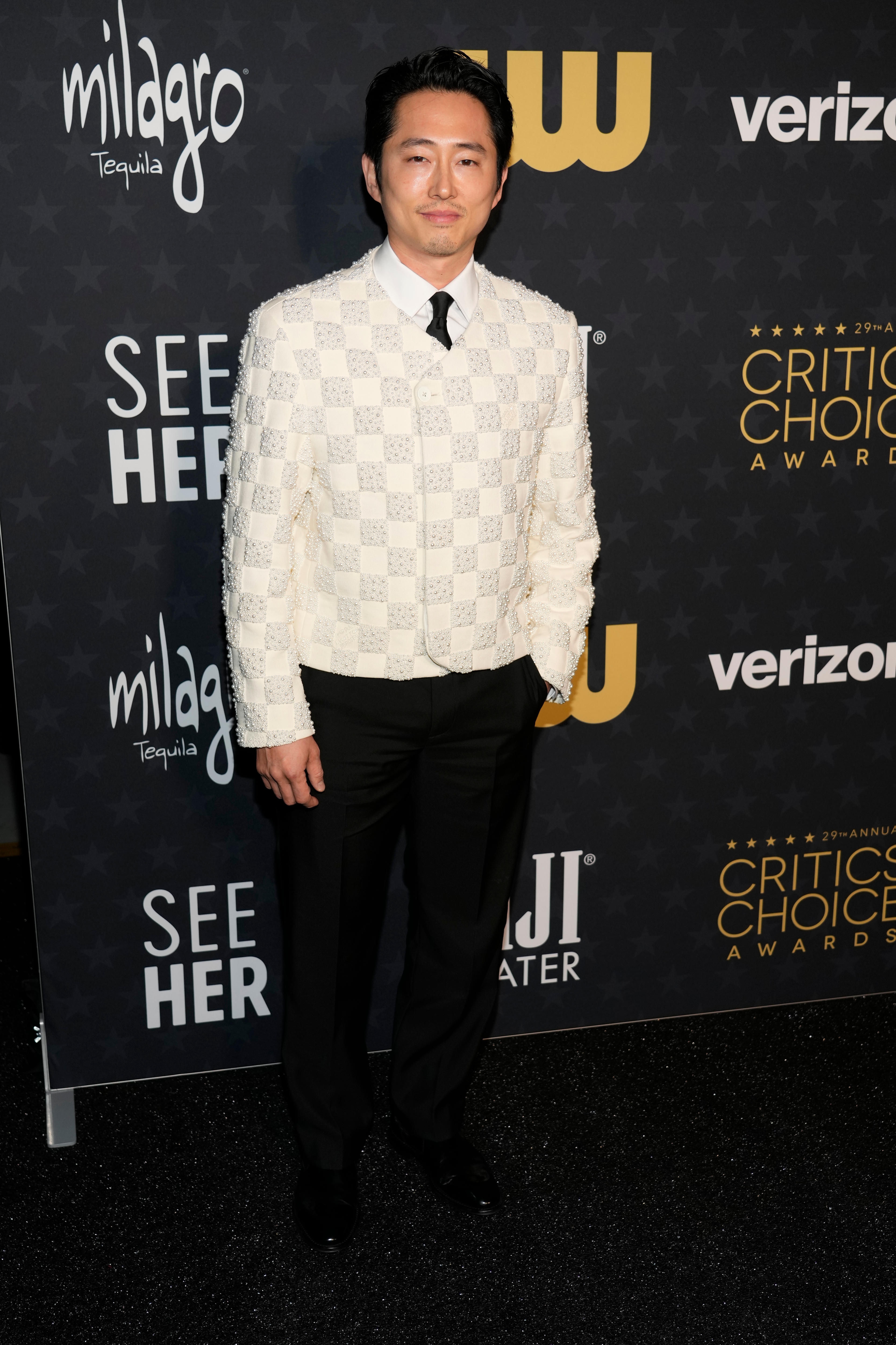 Steven Yeun wearing a white blazers with a white sequinned checker board pattern and black trousers