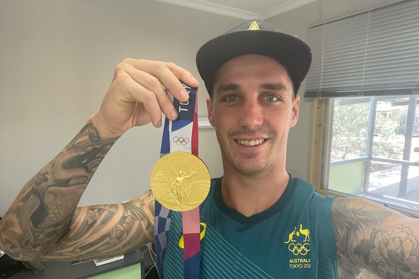 A man with a lot of tattoos in a singlet top and cap holds a gold medal and smiles as he takes a selfie