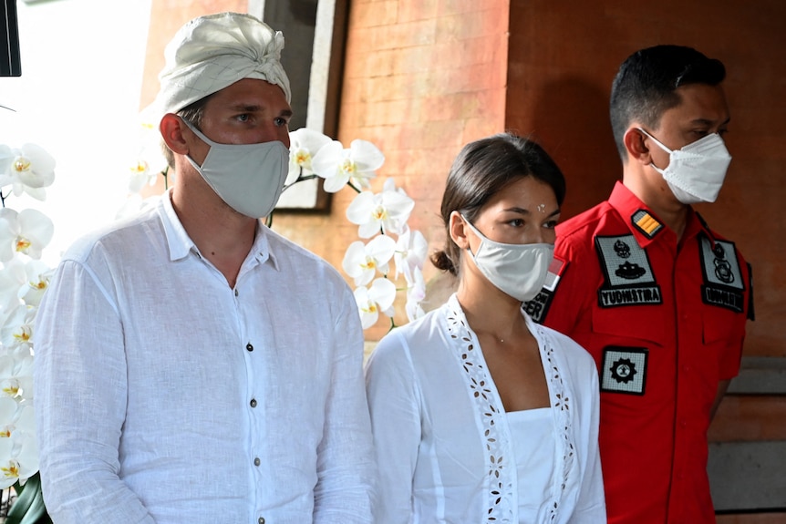 A man in white shirt, and woman in white dress and an official looking man in red suit, all wearing face masks, stare ahead.