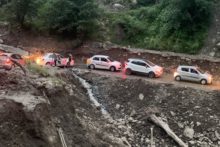 Five cars backed up on a dirt road, a chunk of the mountainside next to them eroded.