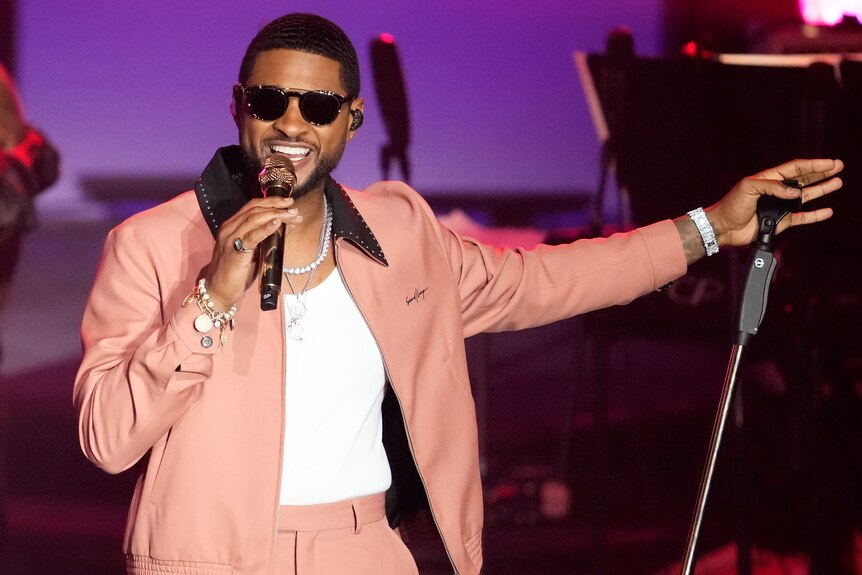 Usher, wearing sunglasses and a light pink jacket and pants, smiles as he sings into a microphone on stage.