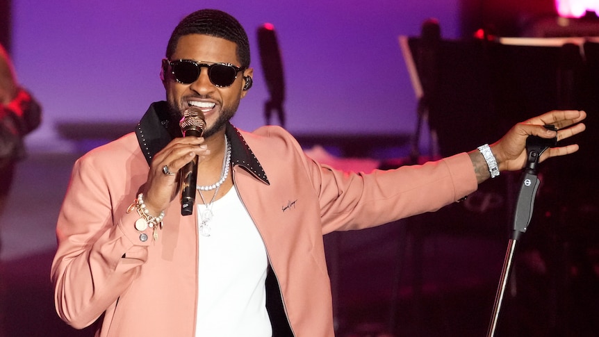 Usher, wearing sunglasses and a light pink jacket and pants, smiles as he sings into a microphone on stage.