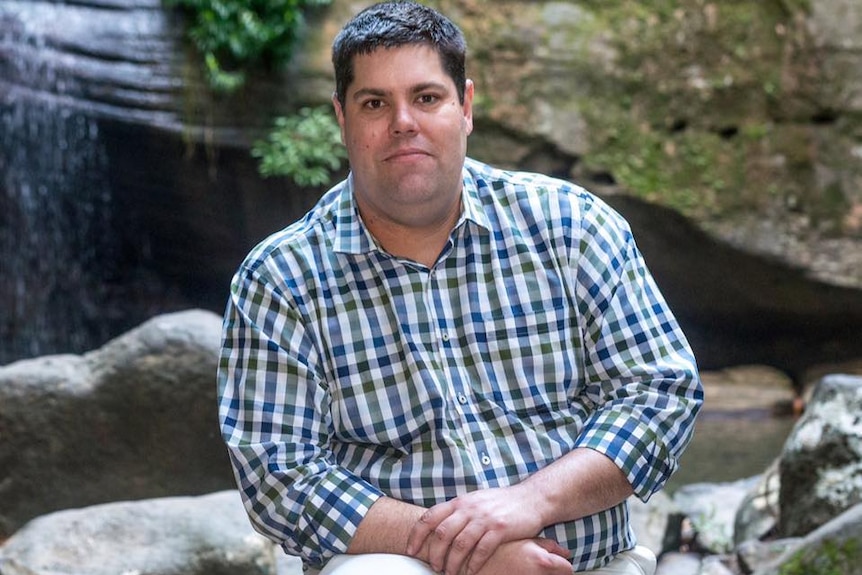 A dark-haired man sits in front of a waterfall.