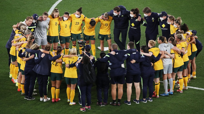 The entire Matildas squad, including coaches, stands in a big huddle in the centre of the ground