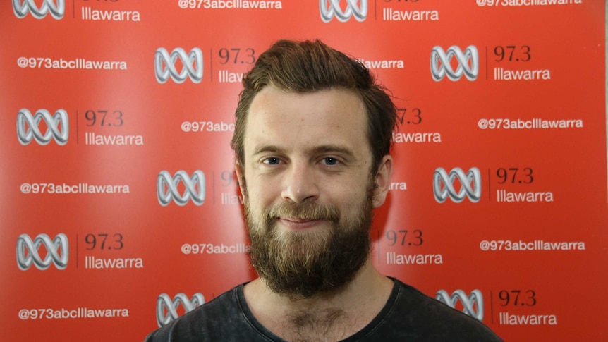 A man with a beard stands in front the ABC Illawarra banner with a slight smile on his face.