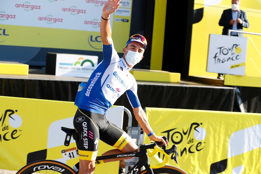 A mask-wearing cyclist waves as sits on his bike in front of the podium at the Tour de France.