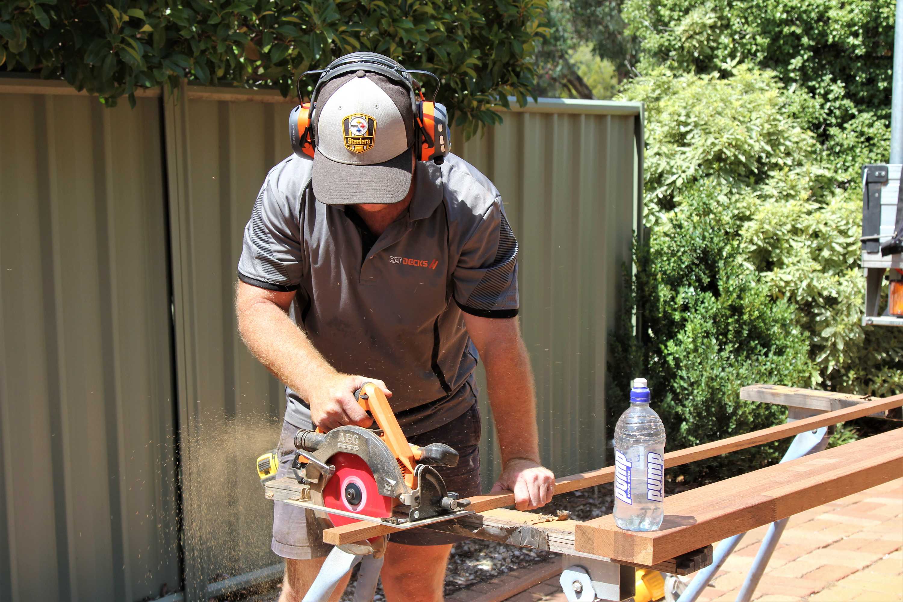 Tradesman cuts wood with saw cutter outside, while water sits on nearby piece of wood.