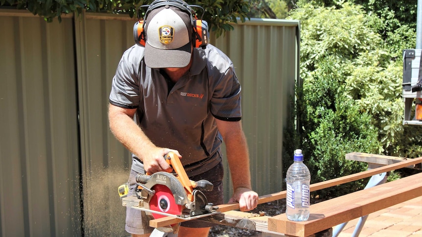 Tradesman cuts wood with saw cutter outside, while water sits on nearby piece of wood.