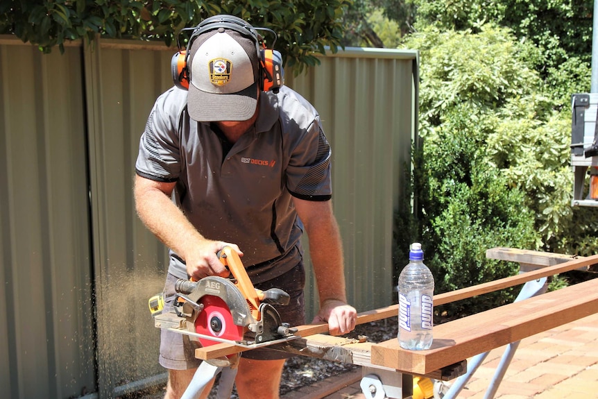 Tradesman cuts wood with saw cutter outside, while water sits on nearby piece of wood.