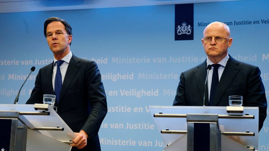 Two men in dark suits speak on semi-transparent glass lecterns bearing the logo of the Kingdom of the Netherlands.