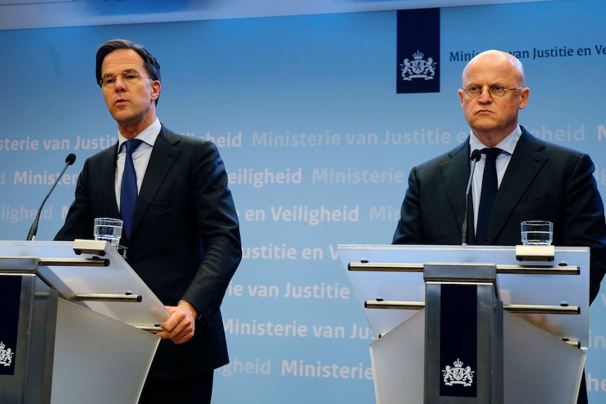 Two men in dark suits speak on semi-transparent glass lecterns bearing the logo of the Kingdom of the Netherlands.