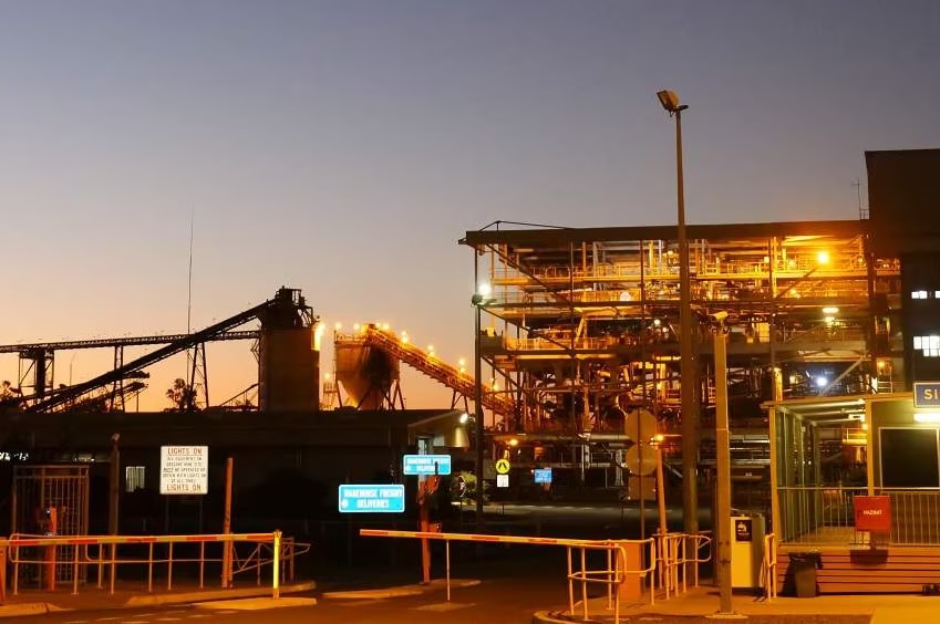 A several-storey high refining set-up at a mine at sunset, with orange lights illuminating stacked machinery. 