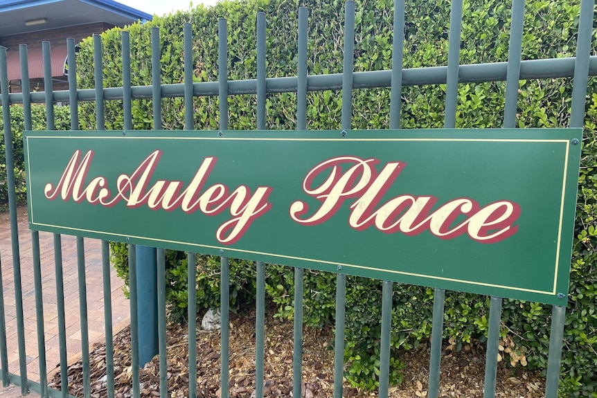 A green sign reads "McAuley Place". It's on a green fence there are green hedges in the background.