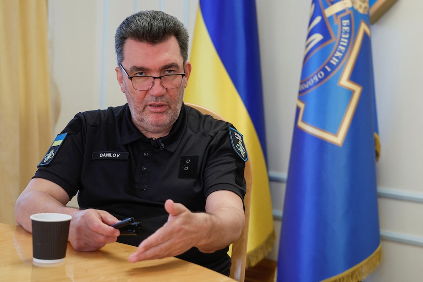 A middle-aged man with grey stubble in a short-sleeved black polo shirt speaks in front of a Ukrainian flag.