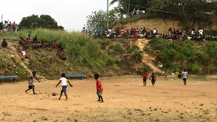 Juniors play soccer on a dirt field in the Solomon Islands, with a big crowd gathering to watch