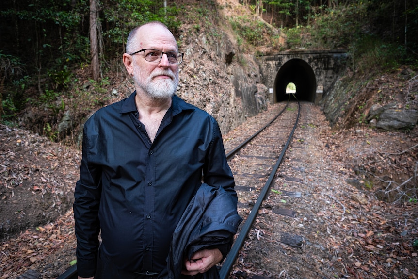 A man wearing a black shirt stands on railway tracks in front of a tunnel.