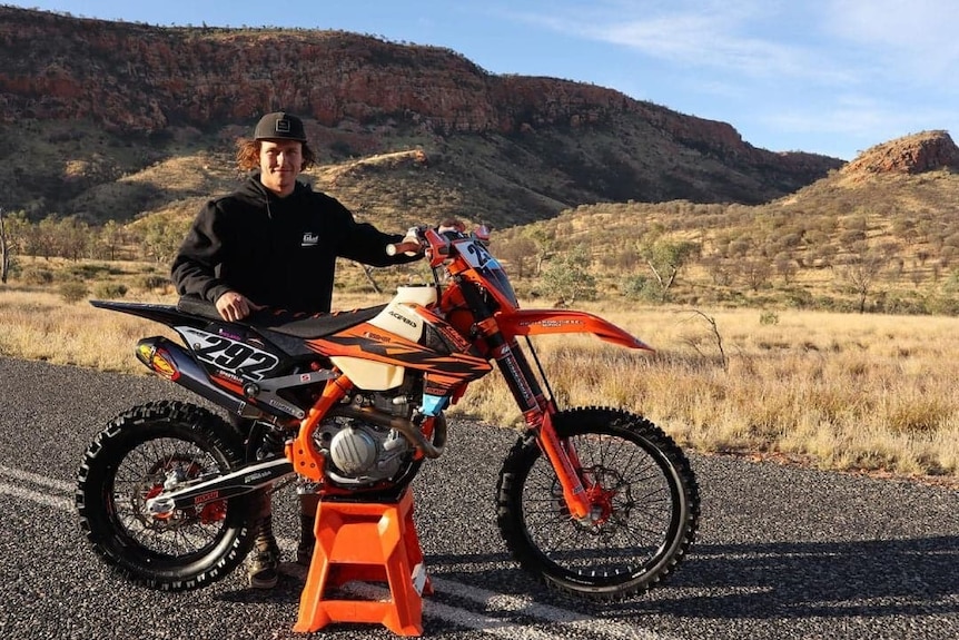 A man wearing a black cap and hoodie stands with his orange dirt bike on a road against the backdrop of the Flinders Ranges.