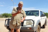 A man in a khaki shirt and shorts standing in front of a white four wheel drive