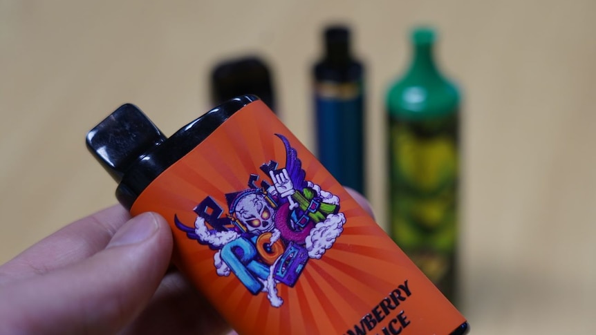 A hand holds a vape containers with some other vapes visible in the background. 