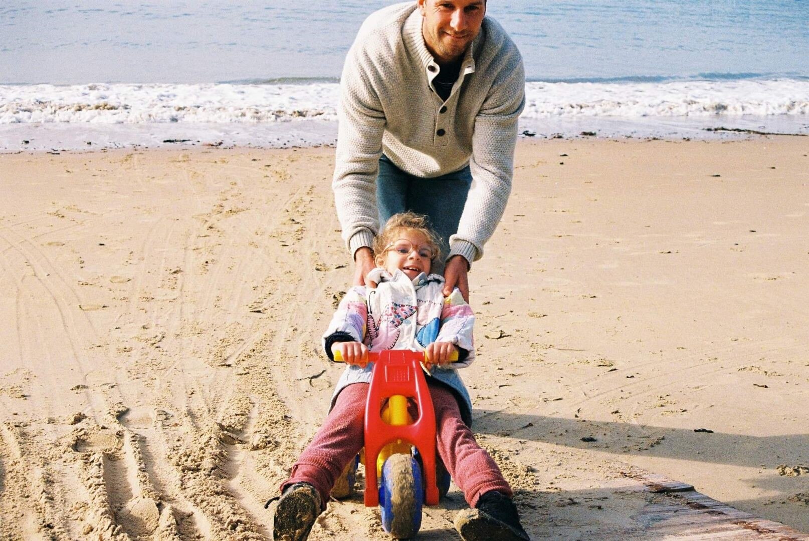 A man on a beach helping his toddler ride a small bike.