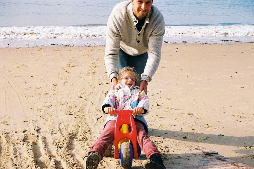 A man on a beach helping his toddler ride a small bike.