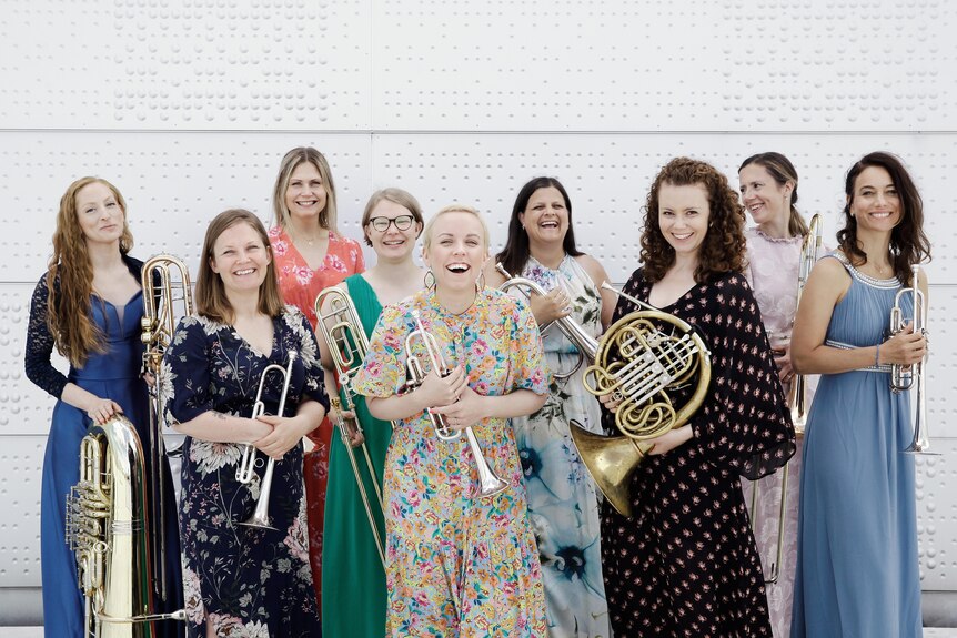 Ten women in brightly coloured dresses stand smiling against a white wall, all holding brass instruments.