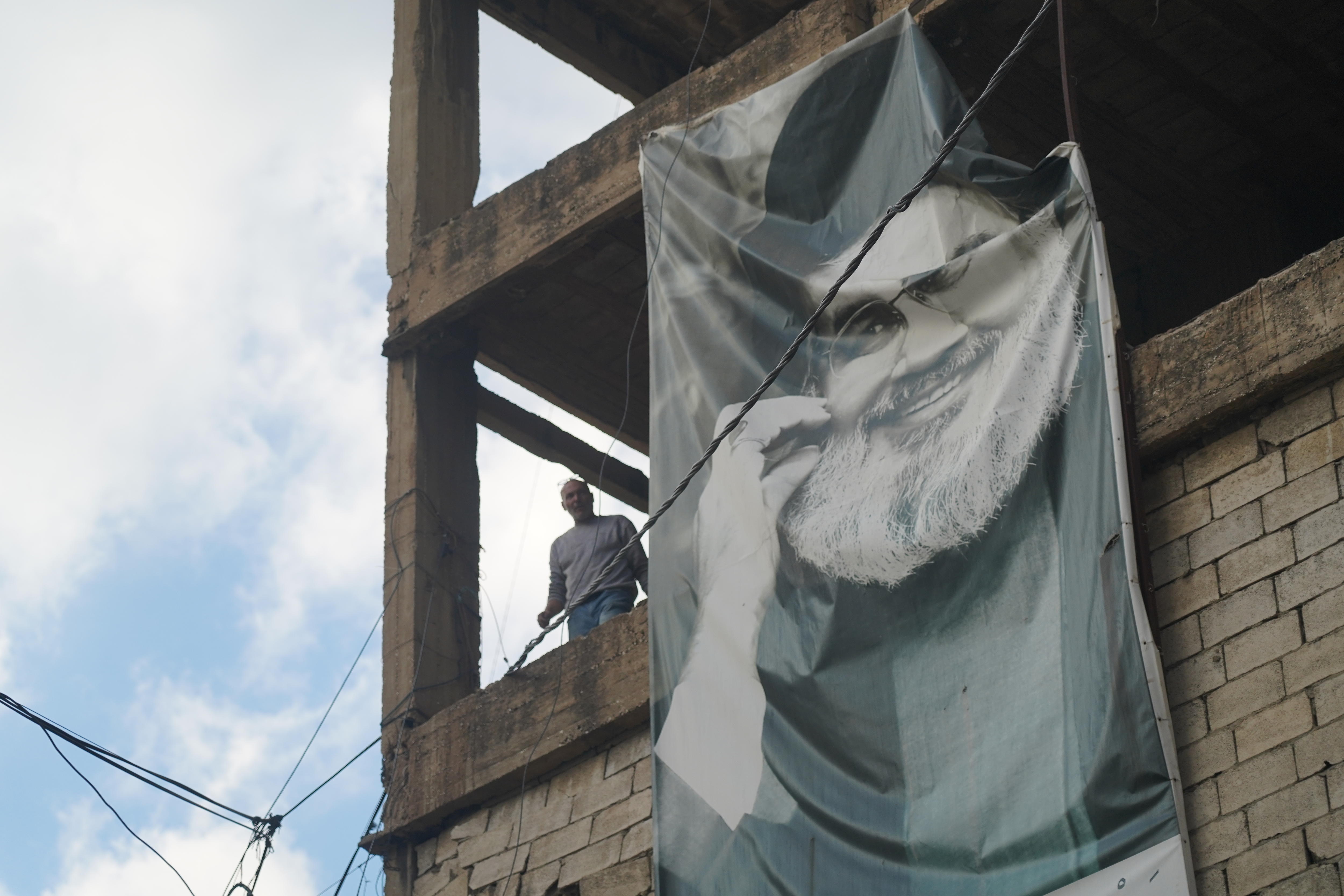 A photo of a man looking out from a damaged building, with a poster of Hassan Nasrallah hanging beside him.