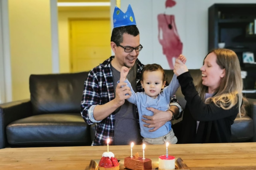 A happy young couple hold up the arms of a toddler who is looking at the candles on a small birthday cake.