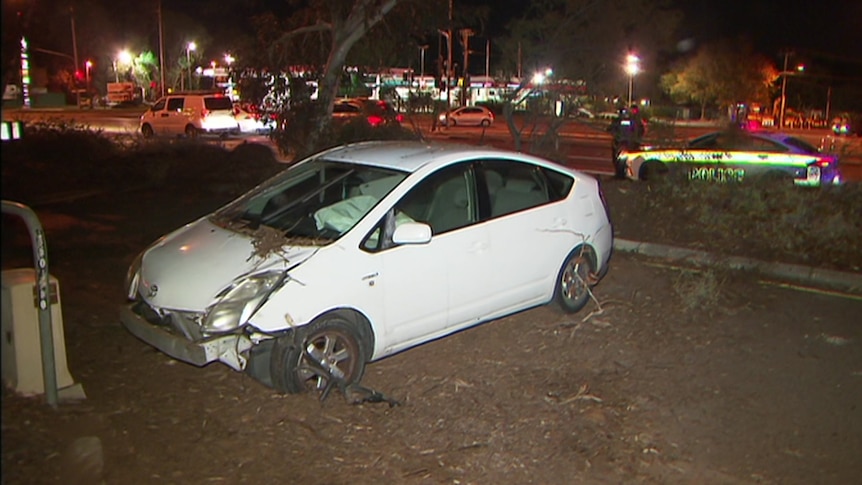 A badly damaged white car in dirt with a police on the road behind